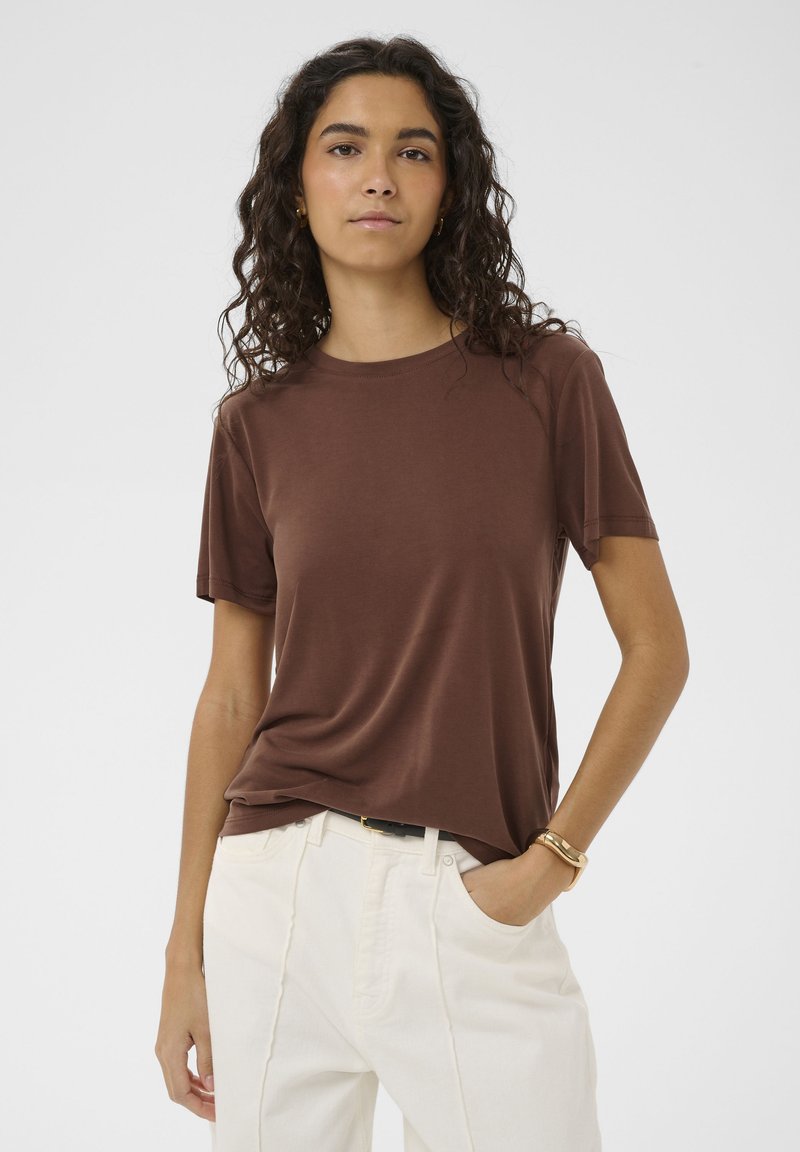 Vrouw met krullend donker haar, gekleed in een bruin shirt met korte mouwen, witte broek en een gouden armband, staande met één hand in haar zak.