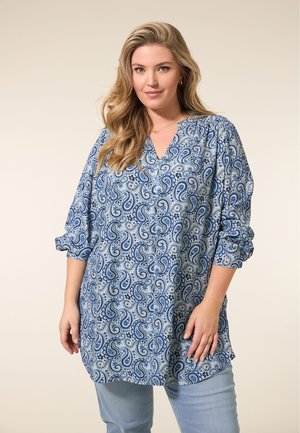 Blauwe paisleypatroon tuniek met een V-hals, driekwartmouwen en geplooide details op de schouders. Gecombineerd met lichtblauwe jeans.