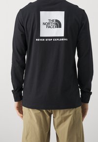 Schwarzes Langarm-T-Shirt mit einem quadratischen Logo auf der Rückseite, darauf steht "THE NORTH FACE" über "NEVER STOP EXPLORING" in weißer Schrift.