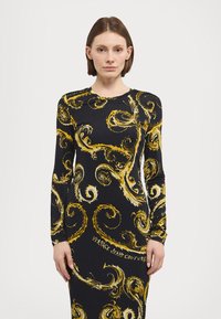Langermet kjole i svart stoff med gullsvirvlende mønstre. Har rund hals og teksten "VERSACE JEANS COUTURE" ved kanten.