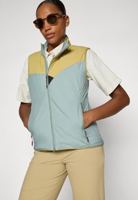 Gilet léger bicolore en vert menthe et jaune avec une fermeture éclair, des poches latérales et un détail en maille noire contrastante. À associer avec un bas beige.