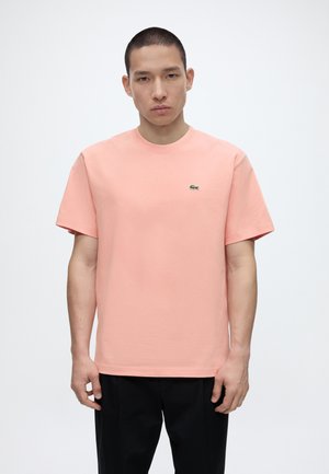 Mężczyzna z krótkimi włosami, ubrany w jednolitą jasnoróżową koszulkę z krótkim rękawem z małym zielonym logo krokodyla na piersi, czarne spodnie.