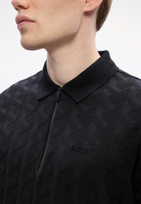 Jauns vīrietis valkā melnu teksturētu polo krekliņu ar rāvējslēdzēja apkakli un diskrētu iepresēto "Boss" logo uz krūtīm.