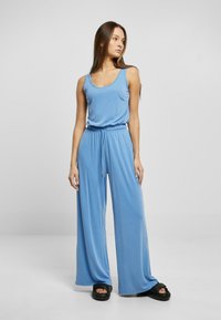 Lys blå jumpsuit med en tettsittende tanktopp og vide bukser. Har en snøring i livet og myk, glatt stoff. Svarte platåsandaler.