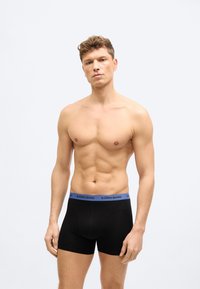 Άνδρας χωρίς πουκάμισο με κοντά σγουρά μαλλιά, φορώντας μαύρα μποξεράκια με μπλε ζώνη μέσης που γράφει "BJÖRN BORG", στέκεται μπροστά σε απλό φόντο.