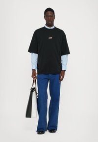 Camiseta negra de gran tamaño superpuesta sobre una camisa azul clara, combinada con jeans acampanados azules. Los accesorios incluyen un bolso tote en blanco y negro.