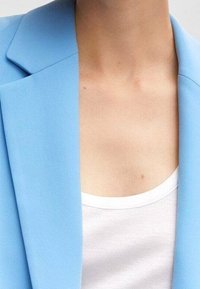 Vaaleansininen bleiseri, jossa on sileä pinta, terävät kaulukset ja istuva hihansuu, puettu valkoisen topin päälle.