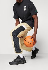 Schwarzes Grafik-T-Shirt mit weißem Design, beige und schwarz farblich unterteilte Jogginghosen und schwarze Sportschuhe mit weißen Socken, während er einen Basketball hält.