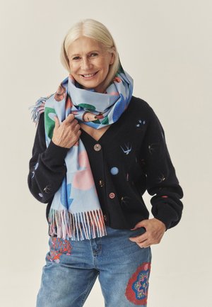 Cardigan negru cu modele brodate, purtat peste o eșarfă albastru deschis cu modele geometrice colorate; blugi din denim cu broderii mixte.