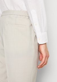 Nahaufnahme einer Person, die beige Hosen mit einer hinteren Paspeltasche und ein weißes Langarmhemd trägt, die Hand entspannt an der Seite.
