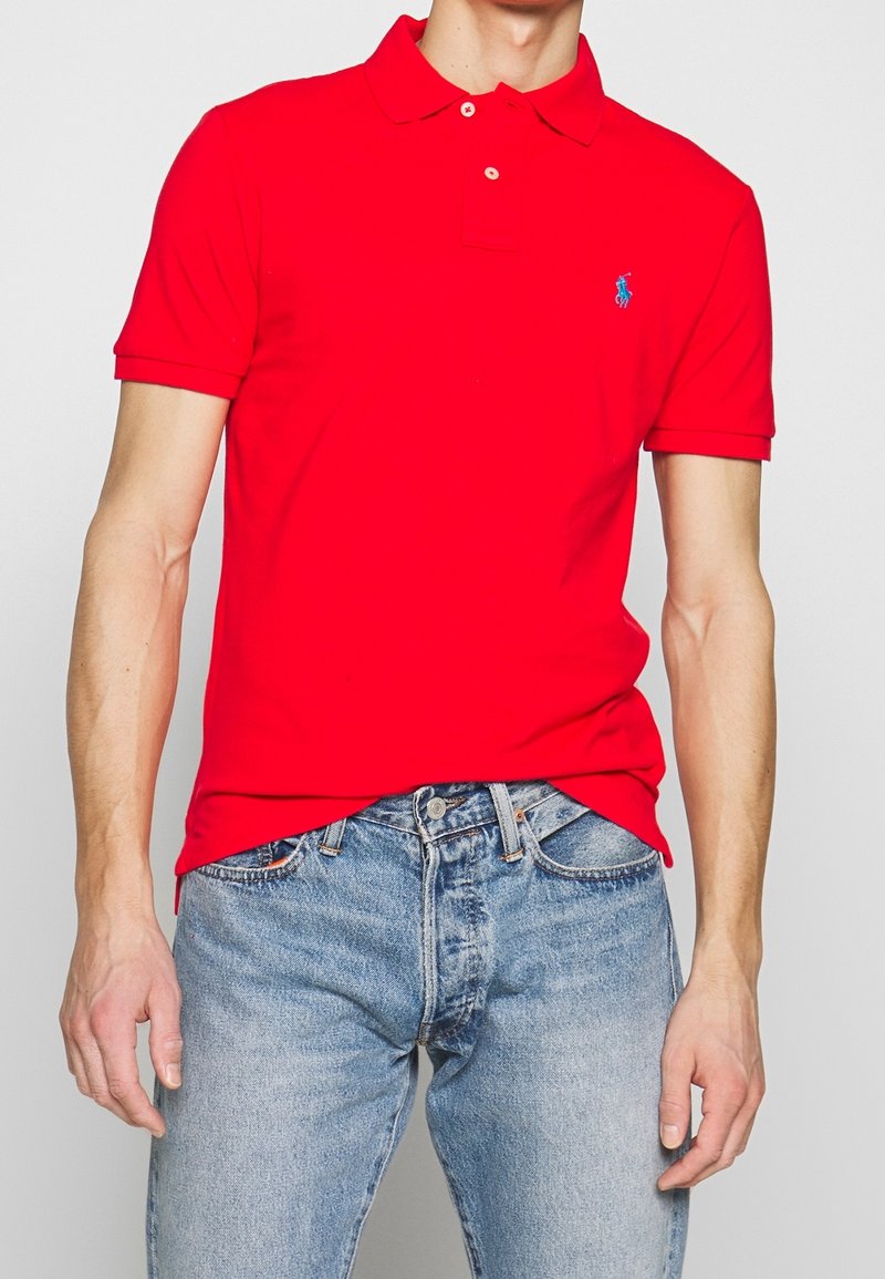 Homme portant un polo rouge vif à manches courtes avec un logo bleu et un jean bleu clair sur fond uni.