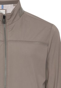 Ljusgrå, zip-up jacka med hög krage, slät textur, med en subtil sömdekoration över bröstet och en stilren dragkedja.