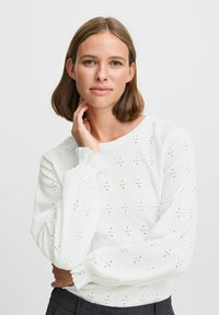 Blusa bianca, con texture e maniche a palloncino, caratterizzata da ricami e collo rotondo. Design semplice e traspirante.