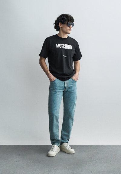 Juoda Moschino marškinėliai su baltu logotipu, šviesiai mėlynos džinsinės kelnės ir balti sportbačiai. Modelis dėvi akinius nuo saulės, kirpčiukai garbanoti ir tamsūs, rankos kišenėse.