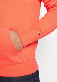 Felpa in corallo arancione con tasca frontale, polsini e orlo a coste. Piccolo etichetta brandizzata sulla manica. Texture liscia.