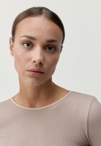 Bežové rebrované tričko s dlhým rukávom a zaobleným výstrihom. Hladká textúra, priliehavý strih a minimálne švy. Neutrálny pozadie zdôrazňuje pozornosť na tričko.