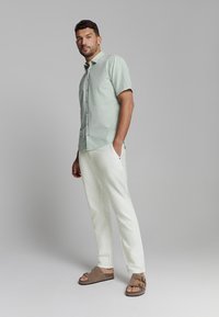 Lichtgroen kortarm shirt met knoopsluiting, gecombineerd met witte broeken en bruine sandalen. Het shirt heeft een subtiele textuur.