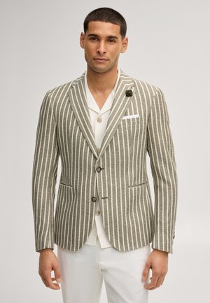Homme portant un blazer beige et blanc à rayures verticales avec une épingle noire au revers, chemise blanche et pantalon blanc sur un fond uni.