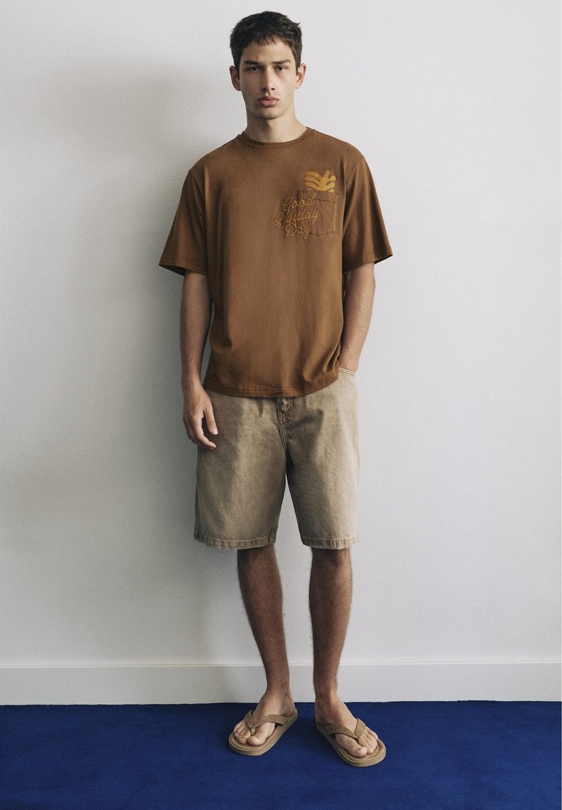 Jeune homme debout contre un mur blanc portant un t-shirt graphique marron, un short beige et des tongs beiges sur un sol bleu.