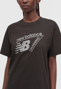 Άτομο που φοράει μαύρο αθλητικό μπλουζάκι New Balance με λογότυπο και κείμενο τυπωμένα ανοιχτό γκρι στο στήθος.