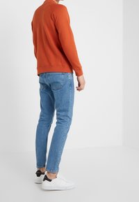 Pomarańczowa bluza, niebieskie obcisłe dżinsy oraz białe trampki z czarnymi akcentami na pięcie. Materiał wydaje się gładki, a krój i design są casualowe.