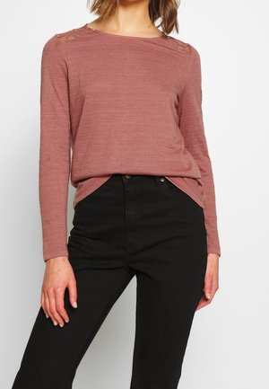 Kobieta ubrana w różową bluzkę z długim rękawem z dzianiny z koronkowymi detalami na ramionach i wysokim stanem czarnych spodni, stojąca na jednolitym tle.