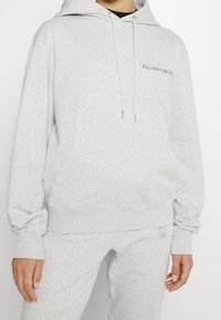 Sudadera gris hecha de una tela suave, con un bolsillo frontal, capucha con cordón y texto bordado en el pecho. Puños y dobladillo acanalados.