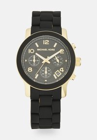 Reloj Michael Kors con correa de silicona negra, caja de acero inoxidable en tono dorado, esfera negra, marcadores de hora dorados y tres subesferas.