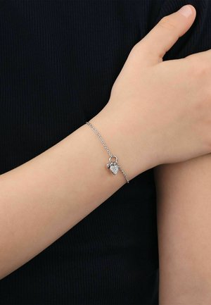 Zartes Silberkettenarmband mit kleinem herzförmigem Anhänger, der auf einem Handgelenk vor einem schwarzen Stoffhintergrund liegt.