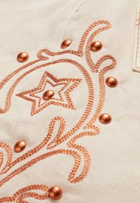 Tela beige con bordados y adornos en color cobre. El diseño incluye una forma de estrella y patrones en espiral con remaches elevados.