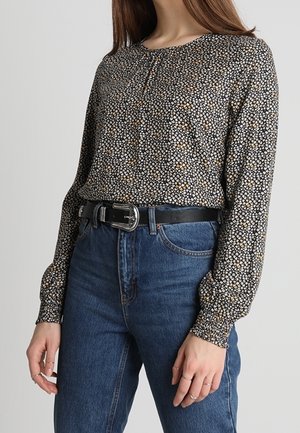 Femme portant un chemisier noir à fleurs à manches longues, rentré dans un jean bleu taille haute avec une ceinture noire et une boucle argentée.
