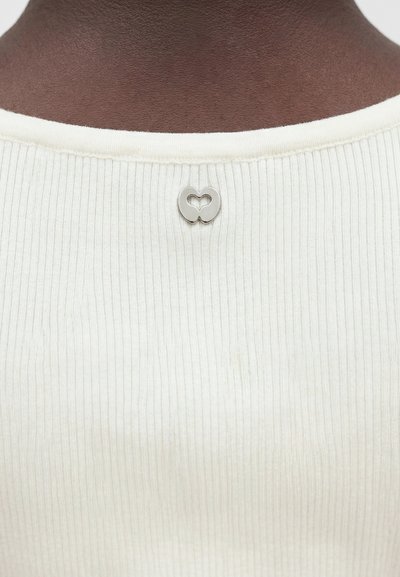 Κοντινό πλάνο ενός σκούρου λαιμού που φοράει ένα λευκό ριγωτό τοπ με μια μικρή ασημένια μεταλλική λεπτομέρεια σε σχήμα καρδιάς στο κέντρο του λαιμού.