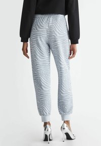 Kvinna i ljusgrå zebra-mönstrade joggbyxor och silverfärgade högklackade skor, stående vänd bortåt mot en neutral bakgrund.