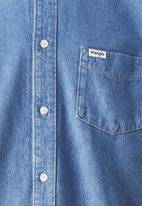 Camisa de denim en azul claro con cuello puntiagudo, botones al frente y un bolsillo en el pecho izquierdo con una etiqueta de "Wrangler". Textura suave.