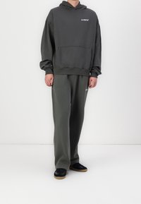 Sweat à capuche gris avec une poche avant et un logo blanc sur la poitrine, associé à un pantalon de jogging gris assorti, et des baskets noires avec des semelles en gomme.