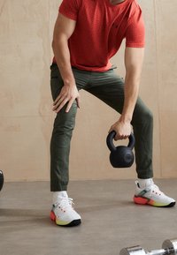 Kettlebell negra etiquetada como "12KG" sostenida en una mano, pantalones gris-verde desgastados, camisa de patrón rojo y zapatos de running coloridos con detalles blancos.