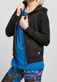 Persona che indossa una felpa nera con zip sopra una maglietta blu, abbinata a leggings con motivo a pixel blu e viola e inserti in rete nera.