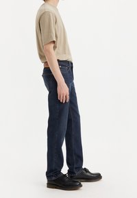 Beige cotton t-shirt, dark indigo jeans, slim fit, denim texture, black lace-up shoes with brown sole, side view of a person.