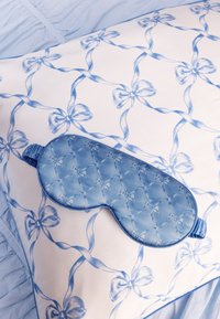 Mascherina per dormire in raso blu con trama trapuntata, posata su una federa bianca con motivo di fiocchi azzurri, su una biancheria da letto azzurra e morbida.