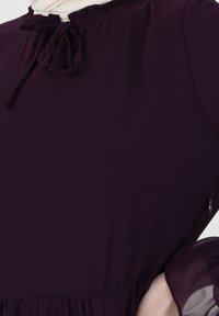 Robe violet foncé avec un col froncé et un détail nœud, manches bouffantes transparentes et fermeture éclair latérale. Texture de tissu lisse.