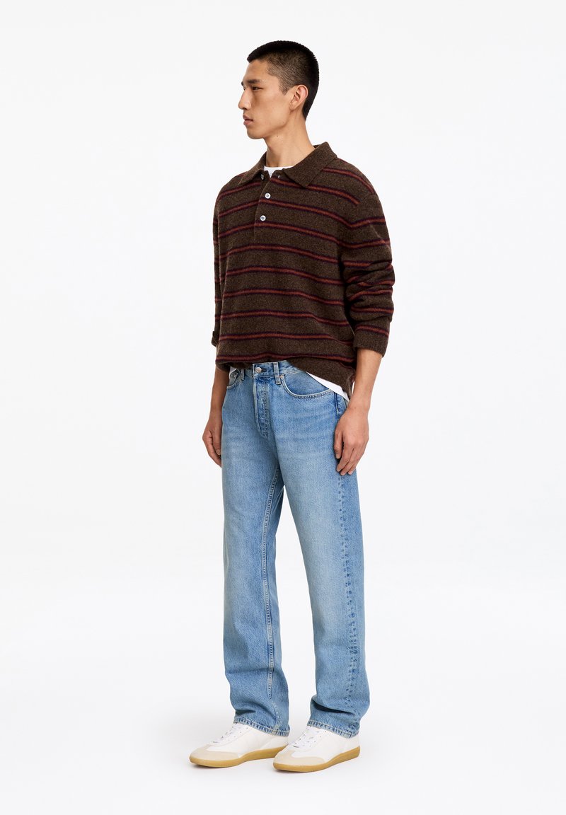 Homme debout portant un polo à manches longues rayé marron et rouge, un jean bleu clair et des baskets blanches sur fond blanc.