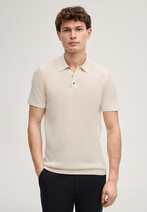 Giovane uomo con capelli corti e ricci che indossa una polo beige a maniche corte e pantaloni scuri, in piedi contro uno sfondo chiaro e semplice.