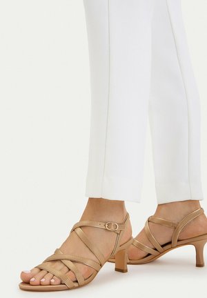 Piedi che indossano sandali beige con cinturini e tacco medio abbinati a pantaloni bianchi aderenti alla caviglia su uno sfondo chiaro.