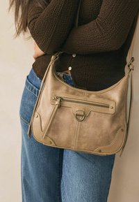 Borsa a tracolla beige in pelle con una tasca frontale con zip, dettagli in hardware dorato e cuciture a contrasto. Abbinata a jeans blu.