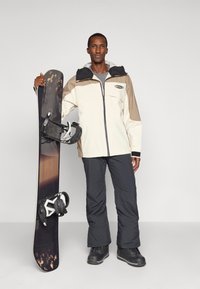 Homem de jaqueta bege e marrom, calças cinzentas e botas pretas, está a segurar uma prancha de snowboard com gradiente preto e castanho e ligações brancas.