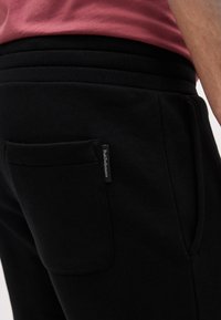 Gros plan sur un pantalon de survêtement noir avec une poche arrière et une étiquette Peak Performance, porté avec un t-shirt rose.