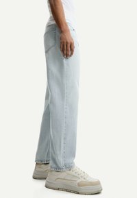 Jeanși din denim bleu deschis, cu croială relaxată, picior drept și buzunare la spate, asortați cu adidași bej cu talpă groasă.