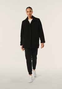 Cappotto nero, textured, di lunghezza media con colletto, tasche laterali, indossato sopra una maglietta bianca, abbinato a jogging neri e sneakers chiare.