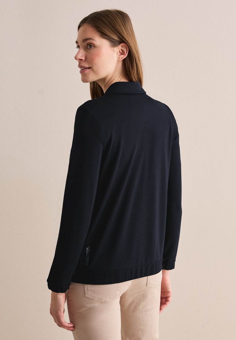 Schwarzes Langarmshirt mit Kragen, glatter Textur, modernem Design und elastischen Bündchen. Sichtbares Markenschild am Saum. Neutraler Hintergrund.