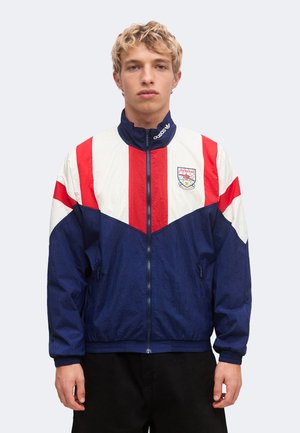 Young man wearing a blue, white, and red zip-up jacket with Arsenal logo and Adidas branding on a light background.